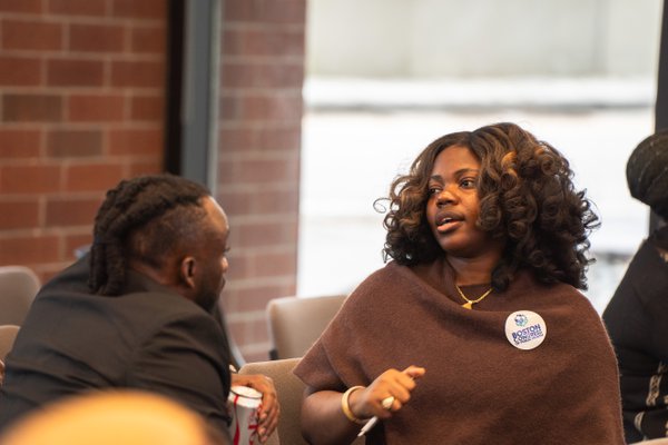 An attendee in a brown wrap, wearing a Boston Congress of Public Health button, mid-conversation with a colleague.