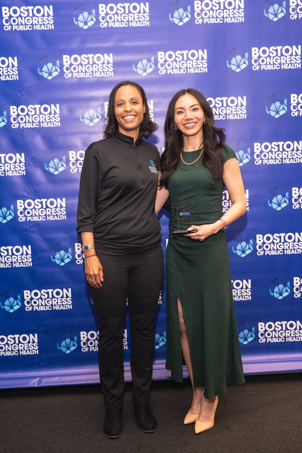 Dr. Candice Carpenter in a BCPH-branded zip top stands beside Dr. Monica Wang in a green dress holding her award, both smiling on the BCPH backdrop.