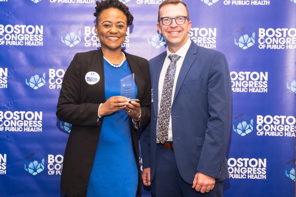 Dr. Juliet Siena Lumati in a royal blue dress holding her award, seated with her mentor, both smiling.