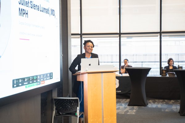 Dr. Juliet Siena Lumati in a royal blue dress, speaking at the podium with her name on the screen behind her.