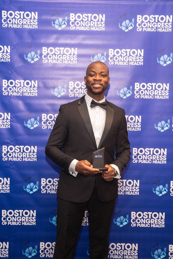 Dr. Ifeanyi Stanley Muoghalu in a black suit and BCPH lapel button, standing solo on the BCPH step-and-repeat.