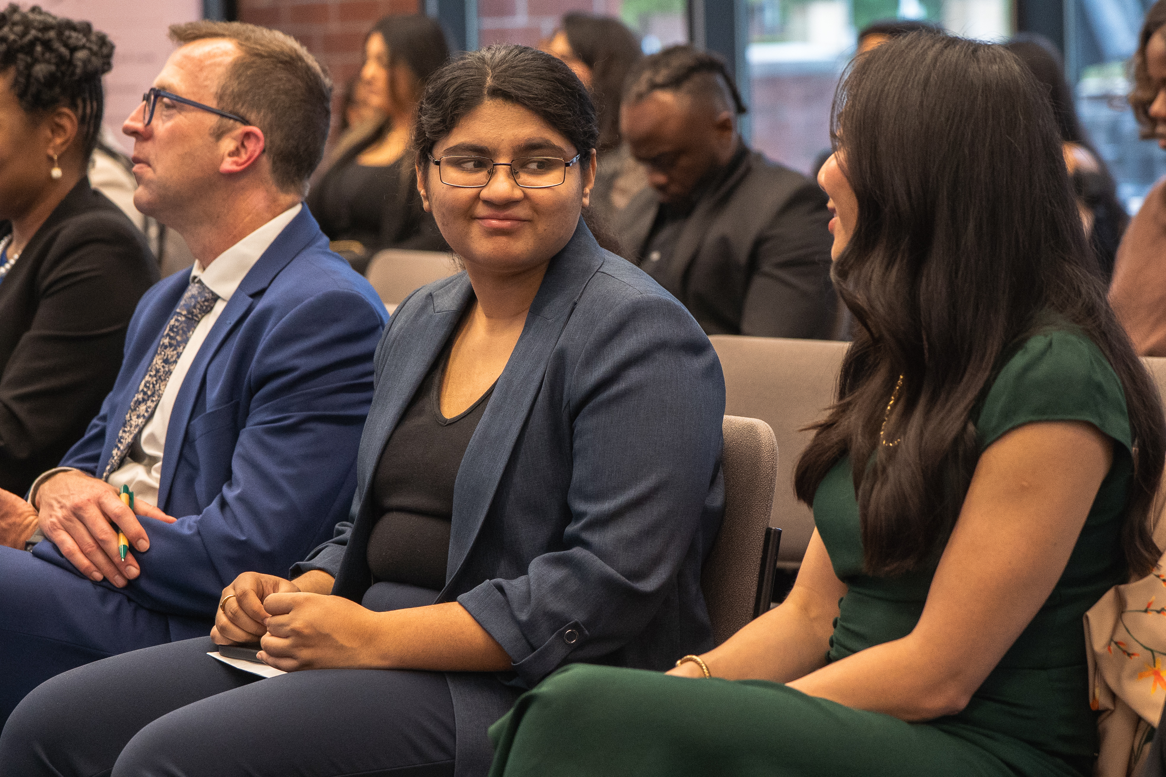 A guest in a blue blazer suit listens attentively in the audience, flanked by two other attendees mid-conversation.