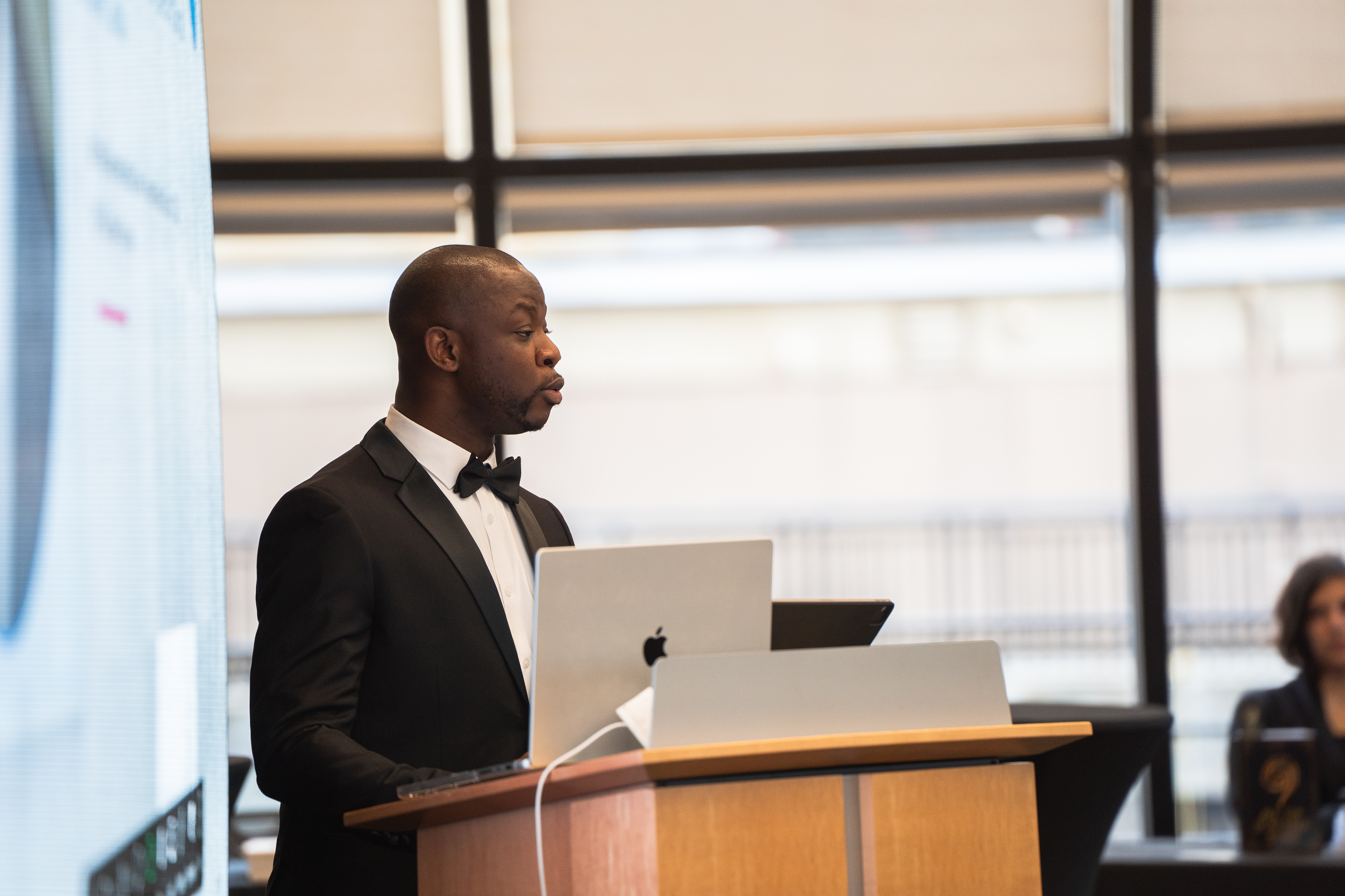 Dr. Olabiyi Olaniran in a black tuxedo with bowtie, delivering the keynote at the lectern.