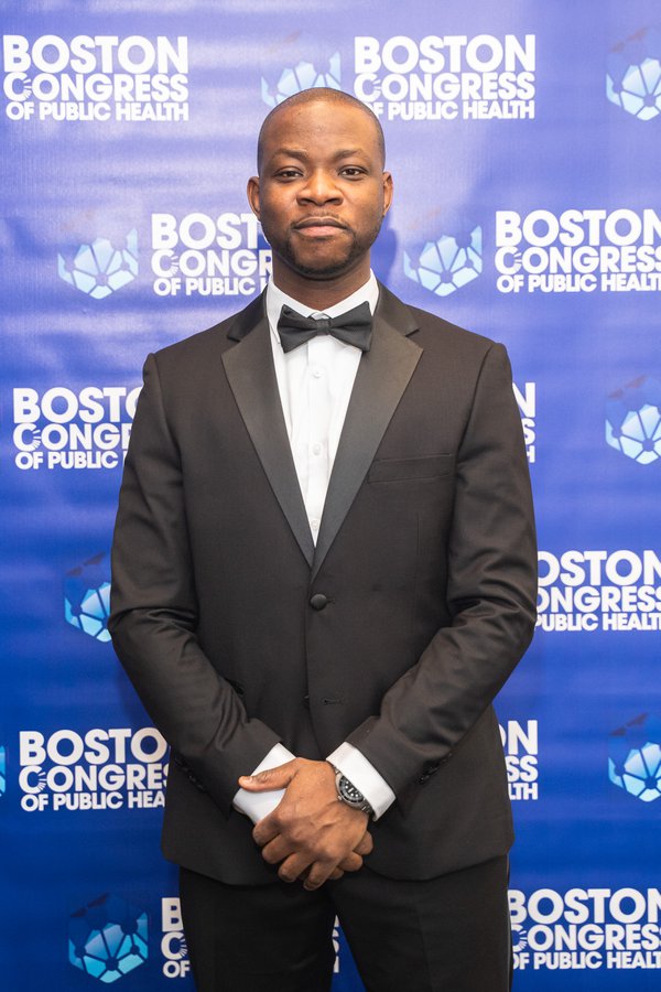 Dr. Olabiyi Olaniran in a black tuxedo with bowtie, standing in front of the Boston Congress of Public Health backdrop.
