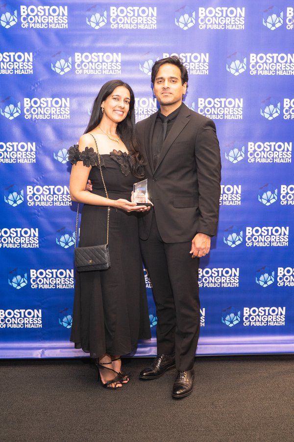 Dr. Kadambari Rawal in a black off-shoulder dress with her companion in a black suit, both holding an award on the BCPH backdrop.