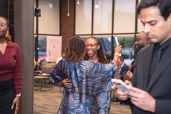 Two attendees embracing at the reception, one in a printed blue wrap, the other smiling over her shoulder.