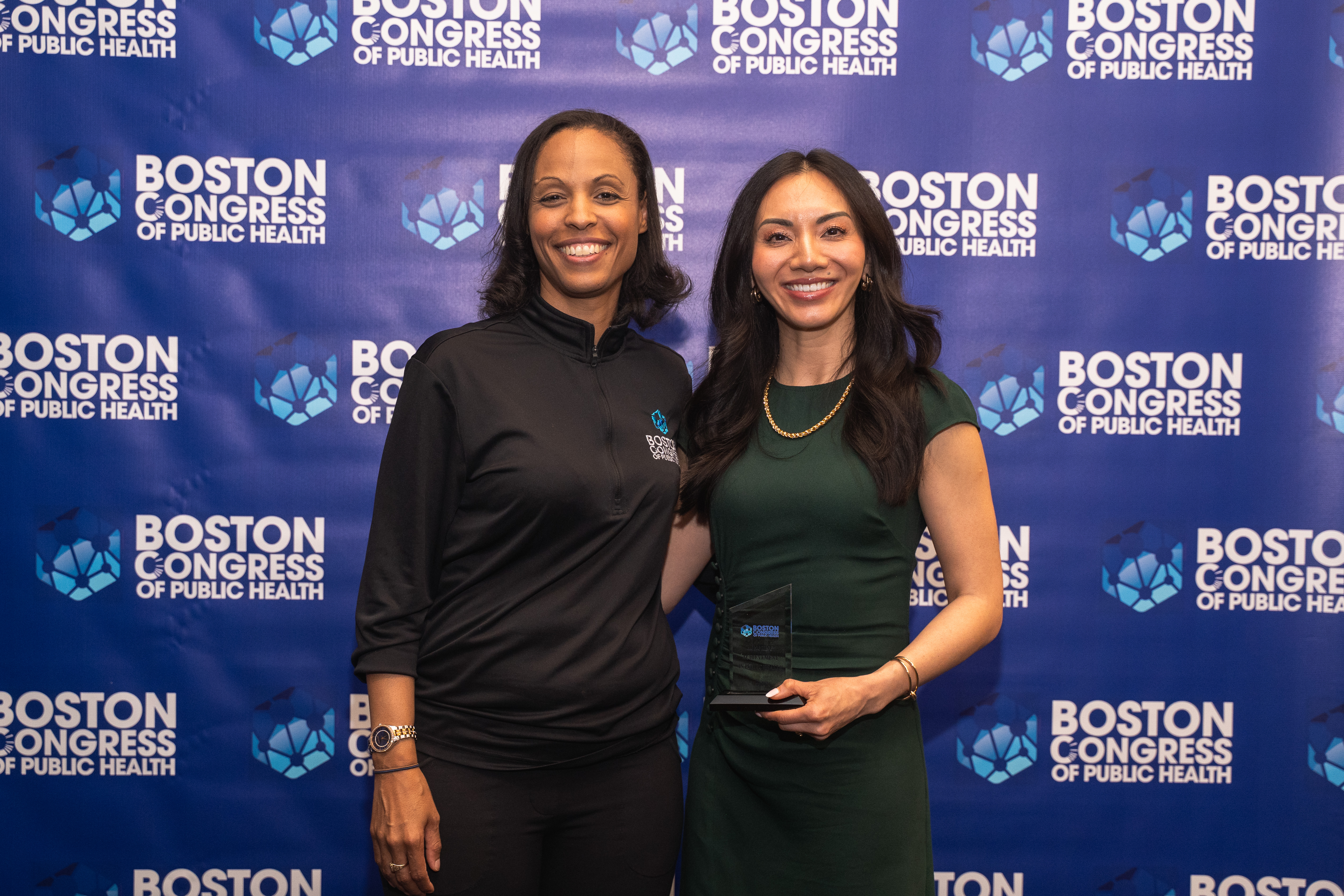 Dr. Candice D. Carpenter in a BCPH-branded zip top beside Dr. Monica Wang in a green dress holding her award on the BCPH backdrop.